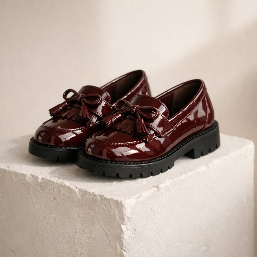 Mocassins fille vernis bordeaux à nœud et franges, posés sur un socle blanc