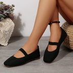 deux ballerines noires portées, jambes croisées près d’un coussin en osier