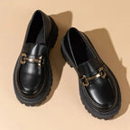 Deux mocassins noirs à mors doré vus du dessus sur fond beige