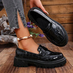 un mocassin vernis noir tenu à la main, l’autre porté, sur plancher bois