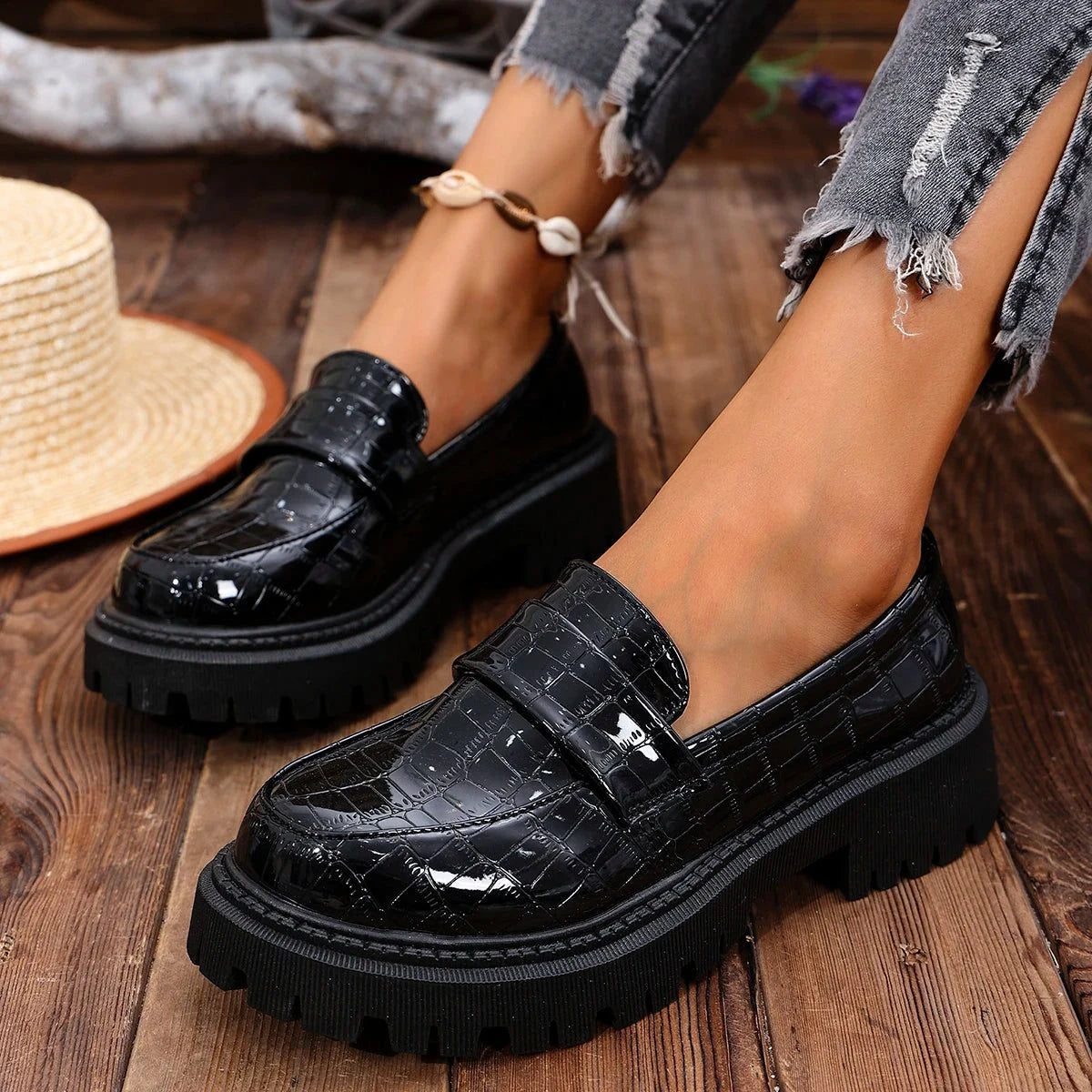 deux mocassins vernis noirs portés, jambes croisées sur un parquet avec chapeau de paille à côté