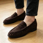 la paire de mocassins marron portée avec un pantalon noir sur parquet