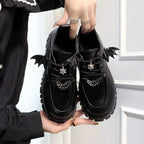 La paire de chaussures noire tenue par une main, détails chauve-souris et chaîne visibles