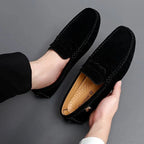 un mocassin noir enfilé, l’autre tenu à la main, fond gris