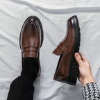 mocassins vernis marron portés, un pied au sol et l’autre tenu à la main