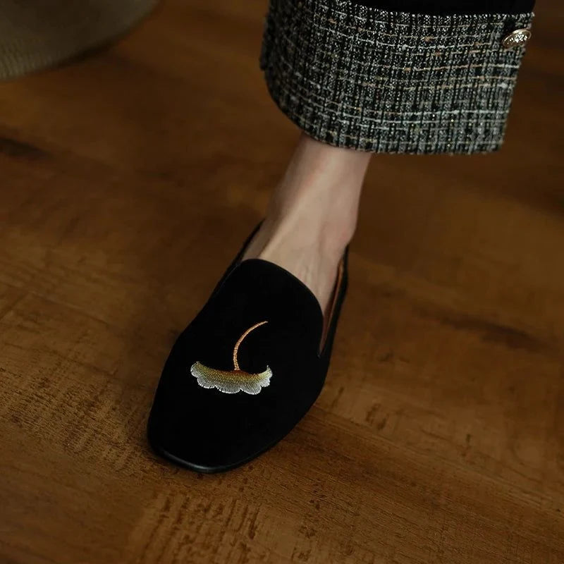 un seul mocassin noir brodé porté sur sol en bois